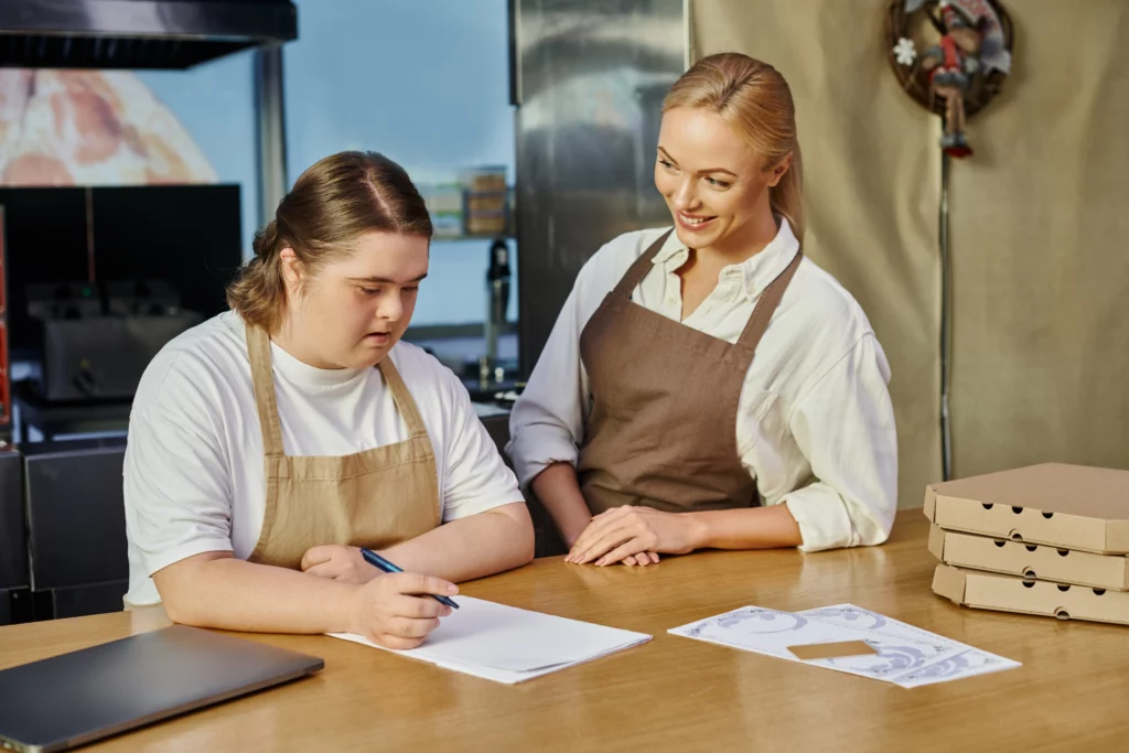 female administrator smiling near employee woman with down syndrome writing order on counter in cafe.