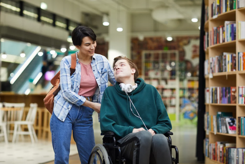 Positive attractive young woman with satchel pushing wheelchair and talking to disabled student in library.